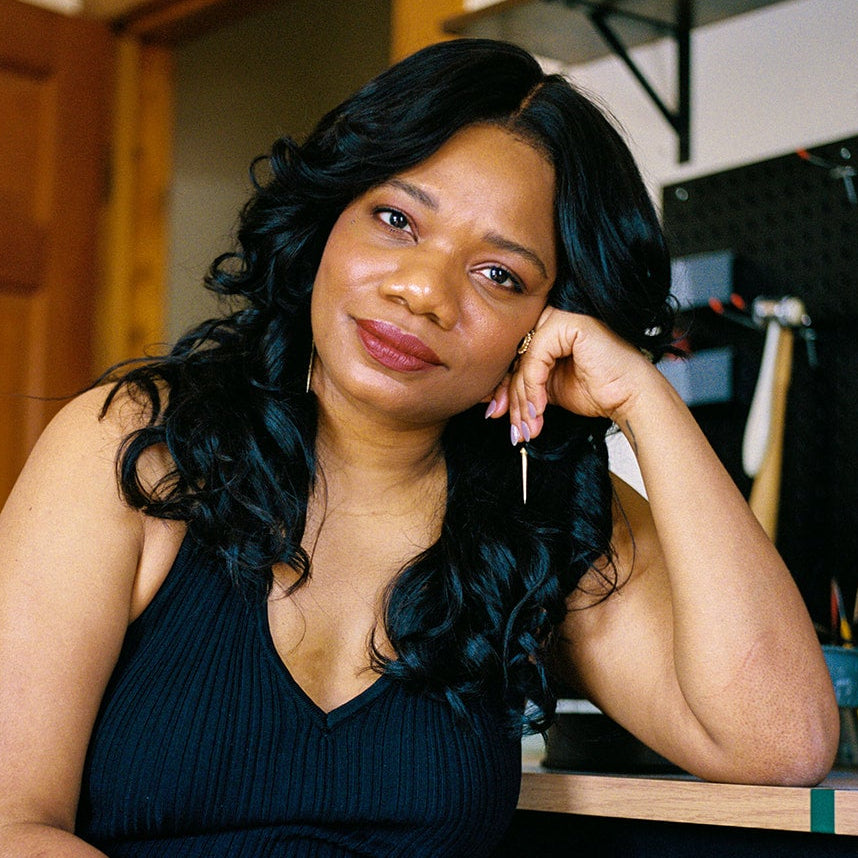 A woman with long, wavy black hair sits in a home office or studio environment, wearing a black ribbed sleeveless top and black skirt. She is leaning on her hand, looking directly at the camera.